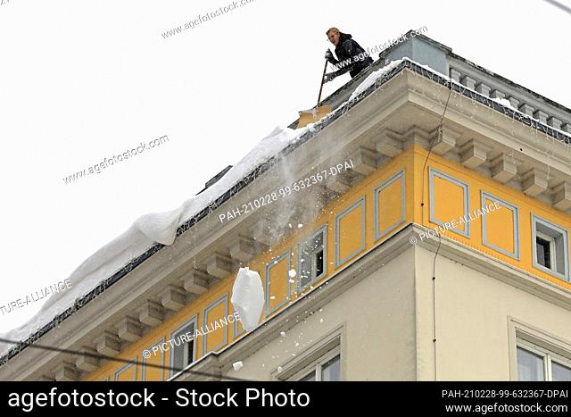 09 February 2021, Saxony-Anhalt, Magdeburg: A man releases overhanging snow slabs from a roof. His colleague secures the footpath