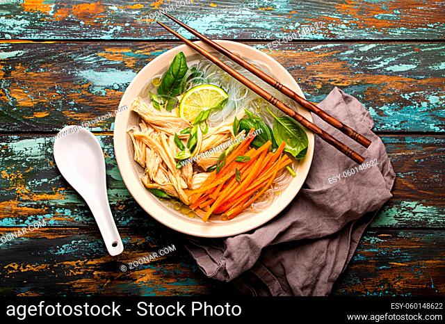 Asian soup with rice noodles, chicken and vegetables in ceramic bowl served with spoon and chopsticks on rustic wooden background from above