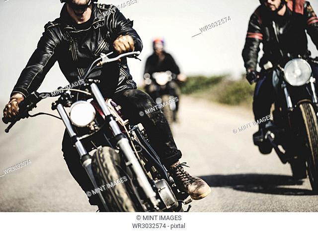 Three men wearing leather jackets riding cafe racer motorcycles along rural road