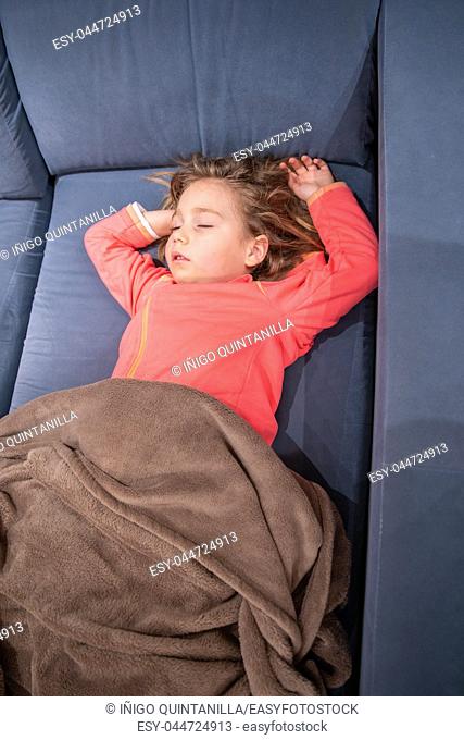 above view of blonde caucasian five years old child, lying on her back on blue sofa, sleeping pleasantly with arms up and covered with a blanket