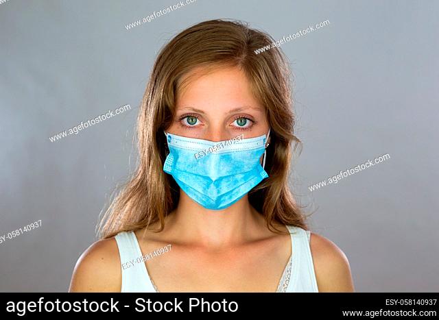 Blonde lady in surgical mask looking to the camera in studio. Young woman with blue respirator during pandemic of coronavirus