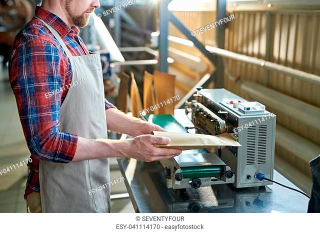 Mid section side view of man wearing apron packing freshly roasted coffee beans in craft paper bags while working in artisan roastery house, copy space