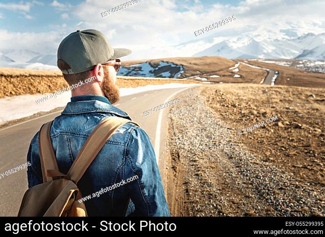 A bearded man in a cap with a backpack ready to go a long way. A man on a country road against the backdrop of mountains and clouds