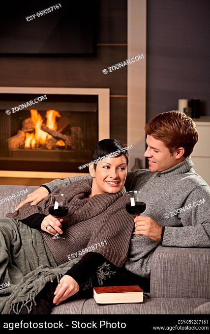 Young romantic couple sitting on sofa in front of fireplace at home, drinking red wine