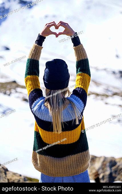 Young woman making heart shape in front of snowcapped mountain
