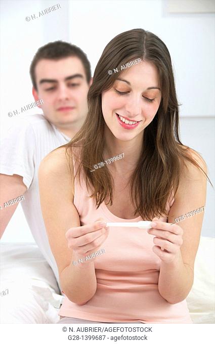 Young couple in bedroom, the woman is smiling and holding a pregnancy test, the young man behind her is waiting for the result
