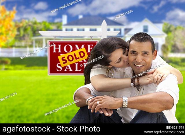 Young happy hispanic young couple in front of their new home and sold for sale real estate sign