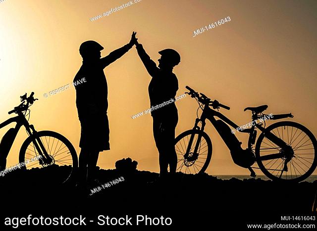 couple of two seniors and mature people together at the sunset with their bikes giving five with the silhouette and the sea at the background