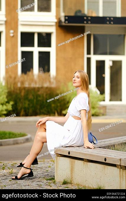 Cute woman in white sundress resting on a bench in residential area