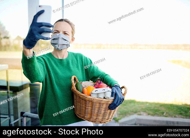 Pretty woman with face mask and green pullover and black gloves makes a phone call after shopping and is holding her shopping basket