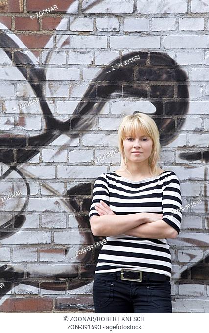 Junge Frau lehnt selbstsicher mit verschränkten Armen an einer Mauer mit Graffito