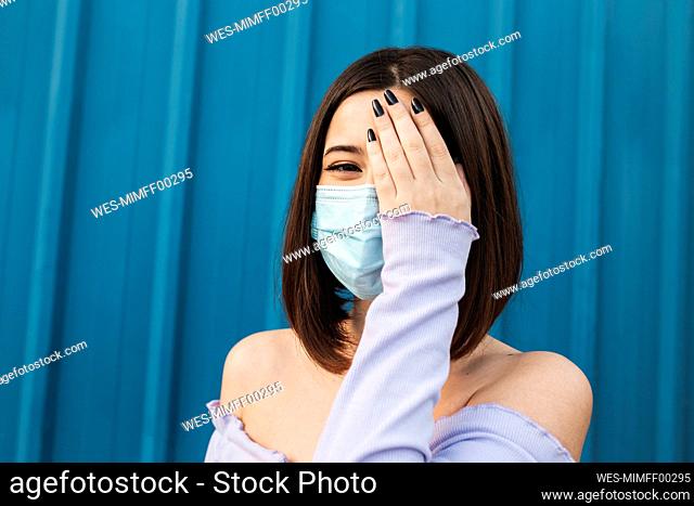 Young woman wearing face mask covering eye with hand while standing against blue wall