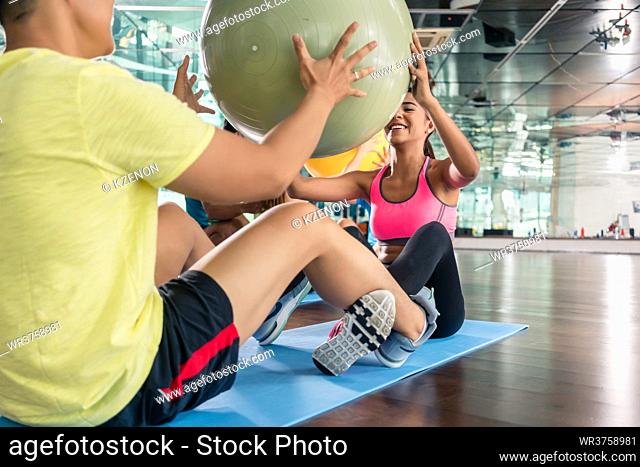 Cheerful young woman giving the fitness ball to her workout partner while doing crunches together for abdominal muscles in a modern health club