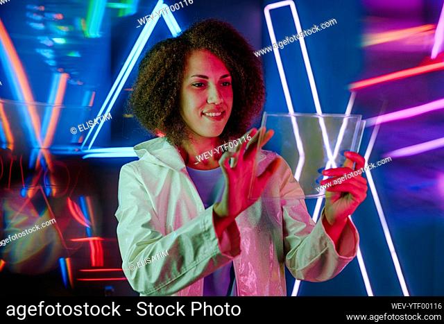 Smiling young woman using transparent laptop in front of neon light