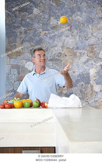 Mature man throwing orange into air standing at kitchen countertop