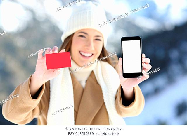 Front view portrait of a woman showing a card and phone screen in a snowy mountain in winter