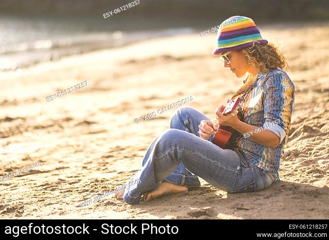 Beautiful woman sitting on sand at beach and playing guitar. Profile of stylish woman in hat and sunglasses playing guitar sitting on beach during holiday