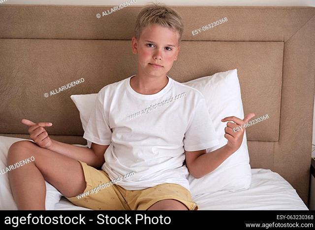 A cute young teenager sits in bed in a white t-shirt and points to his t-shirt. Mockup, copy space
