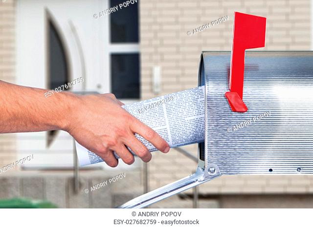 Man removing a newspaper from his mailbox with a closeup of his hand clasping the folded paper against a blue sky