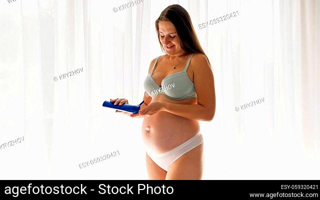 Happy smiling pregnant woman expecting a baby applying moisturizing creme or lotion on her body skin. Concept of beauty, pregnancy and healthcare
