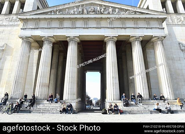 18 February 2021, Bavaria, Munich: Sun-seekers sit on the steps of the Propylaea on Königsplatz. Blue skies and sunshine make the inhabitants of the state...