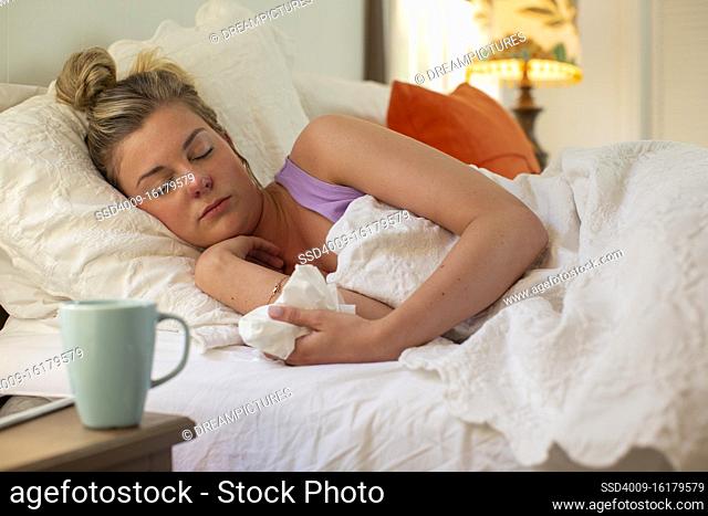 Sleeping young woman in her bed, holding tissue in hand, smartphone and coffee mug sitting on bedside table