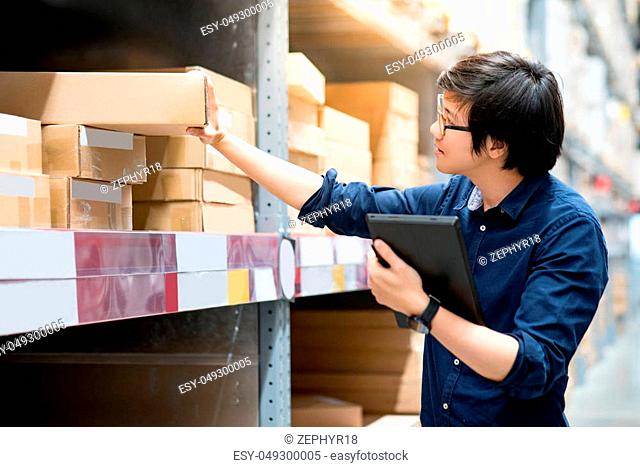 Young Asian man doing stocktaking of product in cardboard box on shelves in warehouse by using digital tablet. physical inventory count concept