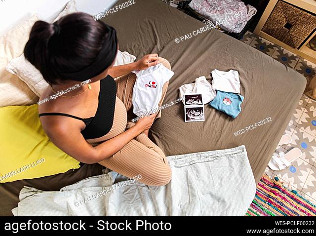 Pregnant woman sitting with x-ray and baby clothes on bed at home