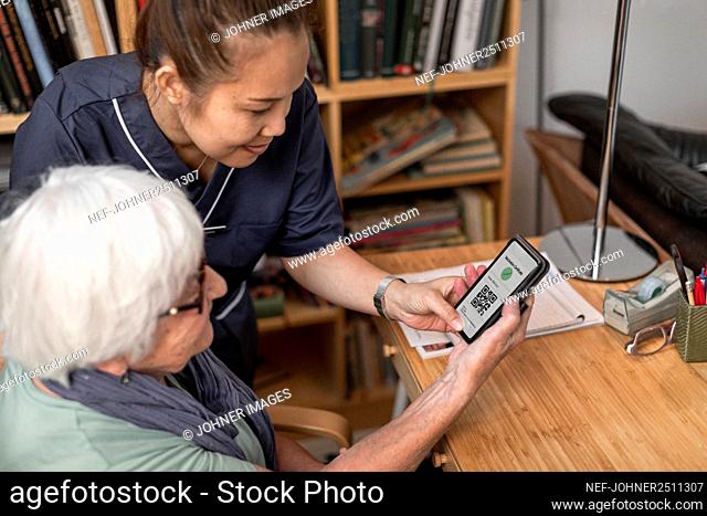 Home carer helping senior woman to use cell phone with Covid-19 vaccine certificate