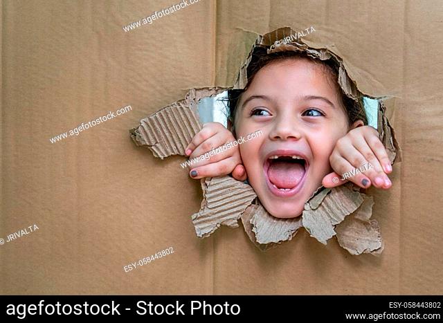 Smiling girl with her mouth open behind a cardboard. The girl is looking through a large hole in the cardboard and with her hands on the edge of the hole