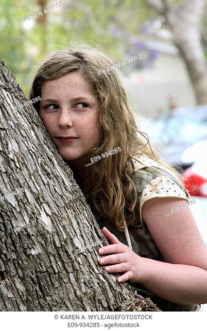 13-year-old girl with blonde-brown long hair and freckles, half leaning against, half hiding behind a tree, up-to-something expression