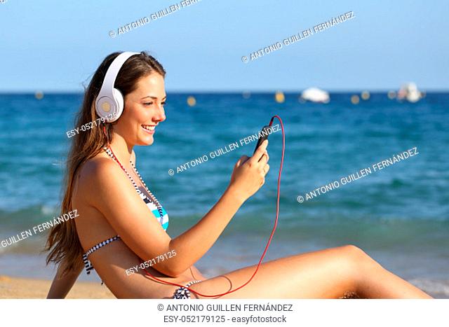 Side view portrait of a happy girl in bikini listening to music using phone and headphones on the beach on summer vacation