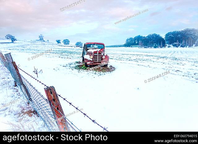 A rusty old vintage car sits in a snow covered field in winter.  There is a leaning rusty barbed wire fence in the foreground .  Space for copy