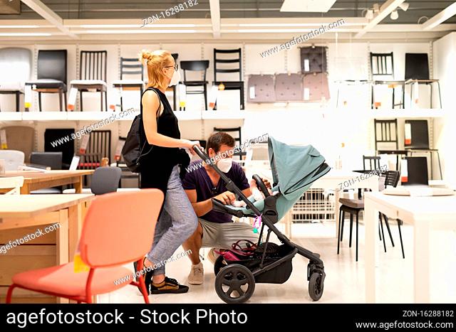 Young family with newborn in stroller shopping at retail furniture and home accessories store wearing protective medical face mask to prevent spreading of...