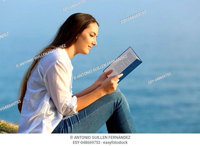 Side view portrait of a relaxed woman reading a book sitting on the beach with the sea in the background