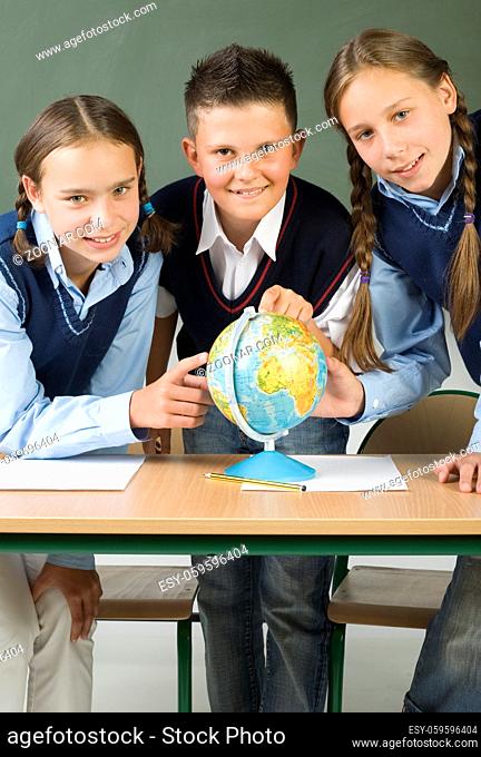 Two, young girls and one boy standing over globe and showing something. Looking at camera, front view