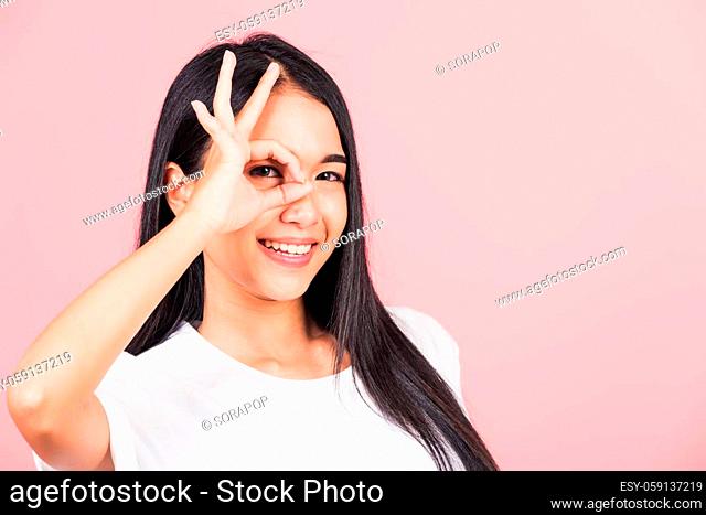 Portrait Asian beautiful young woman standing, She made finger OK symbol sign to agree near eyes looking at camera, studio shot isolated on pink background