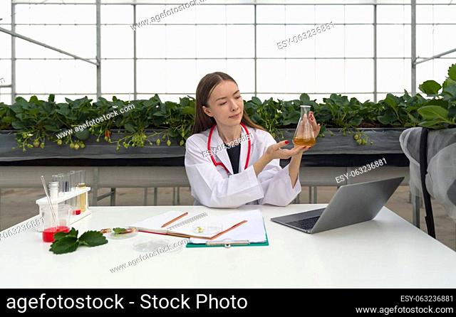 Caucasian female botanical scientist in white gown observes yellow chemical in Erlenmeyer flask. The table is full of research papers, laptop computer