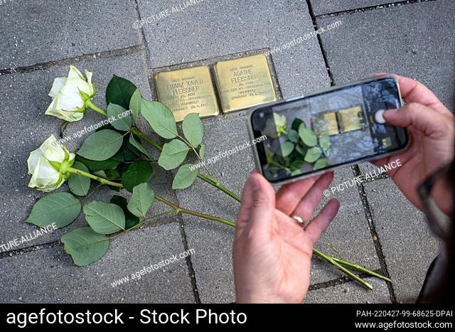 27 April 2022, Bavaria, Penzberg: Roses lie next to two, of the ""Stolpersteine"" made and laid by artist Gunter Demnig, on a sidewalk in front of a house in...