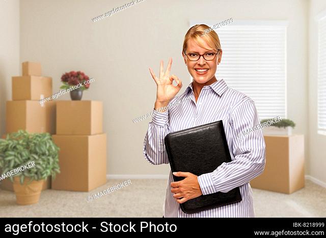 Female real estate agent with okay sign and binder in empty room with packed moving boxes and plants