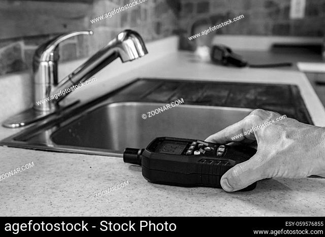 A close-up, black and white view of a person using an indoor air quality monitor, checking for pollutants and allergens inside a residential kitchen