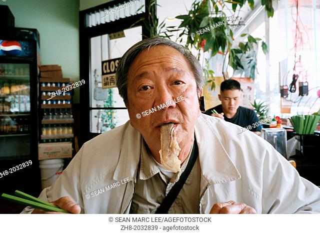 An older Asian man dangles a piece of beef from his mouth like a tongue at a restaurant in San Francisco, California