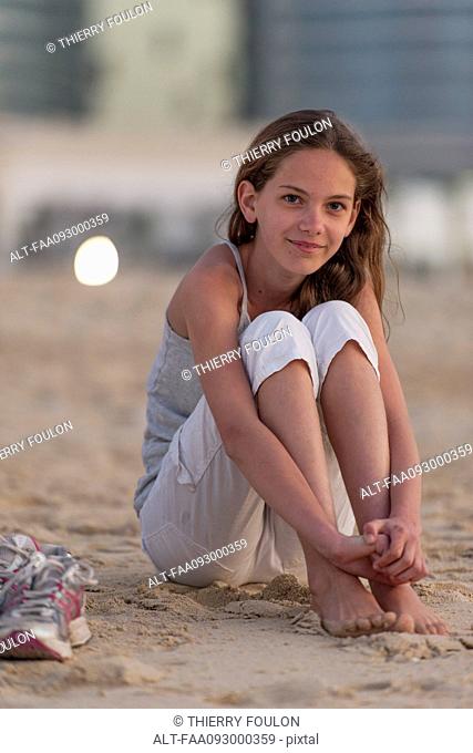 Preteen girl sitting on beach with barefeet, hugging knees