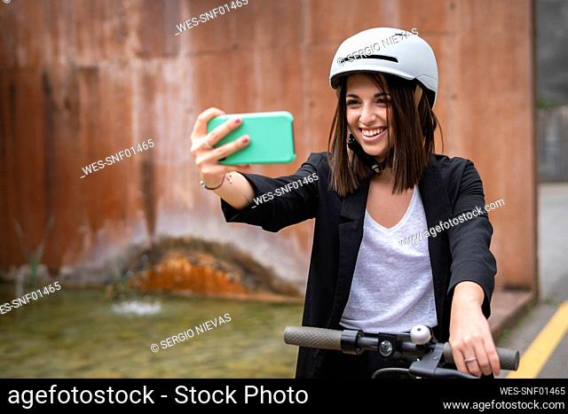 Smiling businesswoman with electric push scooter and cycling helmet taking selfie through smart phone