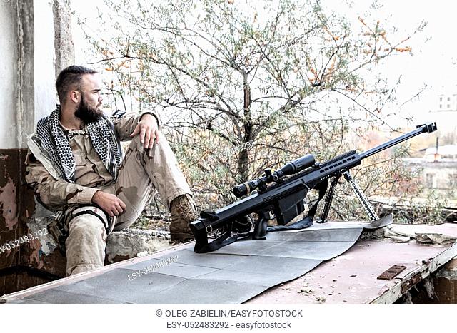 Navy SEAL sniper in battle uniform and shemagh on shoulders, sitting near large caliber sniper rifle with optics sight, on firing position in abandoned building
