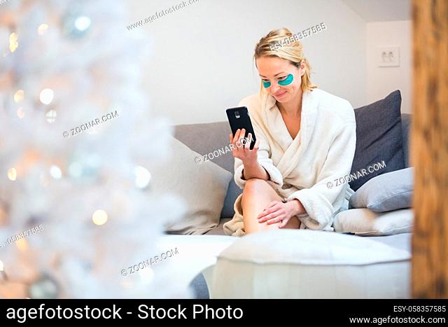 Woman wearing cosy warm bathrobe and cosmetic eye patches relaxing on sofa using social media on phone for video chatting with her loved ones in winter...