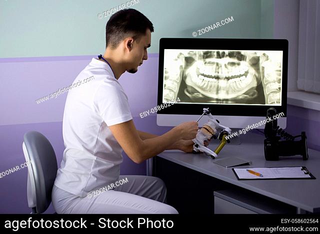 Observant dentist in a white uniform sits on the chair at the table. He looks at an articulator with teeth mould which he holds in his hands