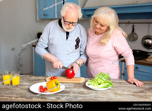 Elderly man and woman spending time togethr cooking. Old man with gray hair cutting red bell pepper with his wife next to him
