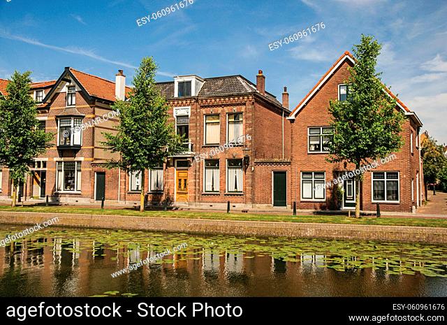 Tree-lined canal with streets on the banks and brick houses on sunny day in Weesp. Quiet and pleasant village full of canals and green near Amsterdam