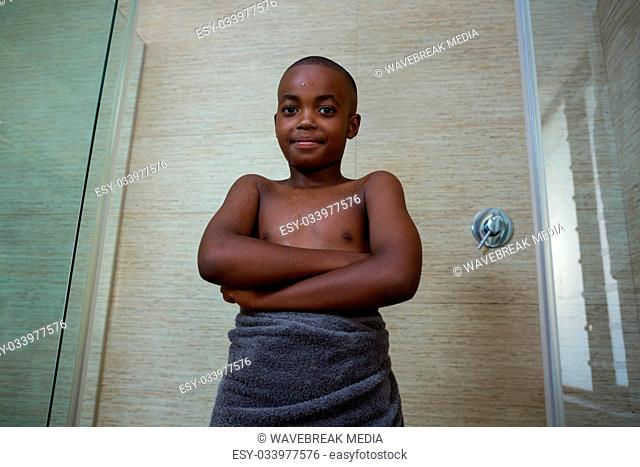 Low angle portrait of shirtless boy wrapped in towel standing with arms crossed amidst glass